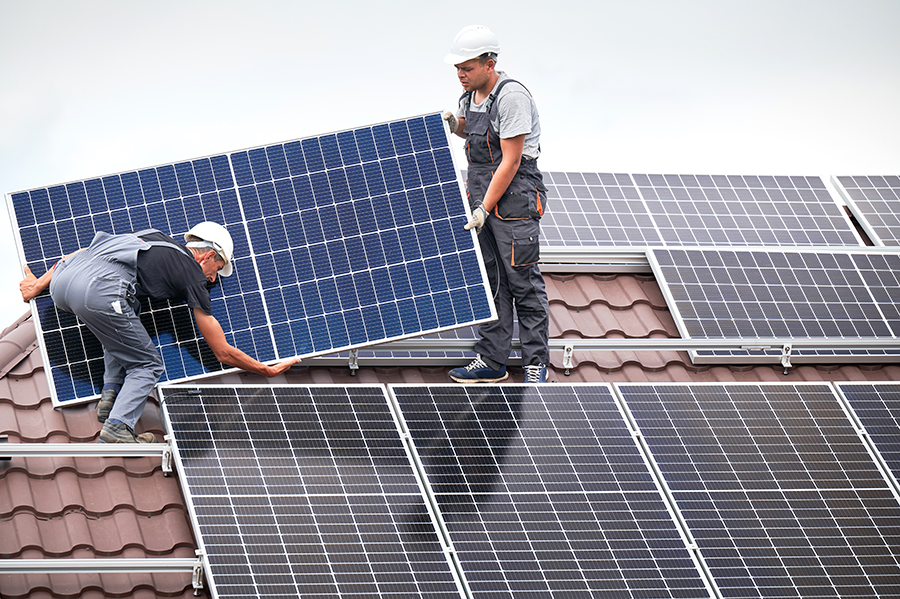 Men technicians carrying photovoltaic solar moduls on roof of house. Engineers in helmets installing solar panel system outdoors. Concept of alternative and renewable energy.